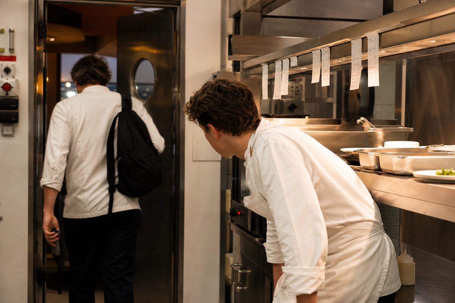 A head chef leaving the pass at the end of a last service, with a younger cook still on the station.